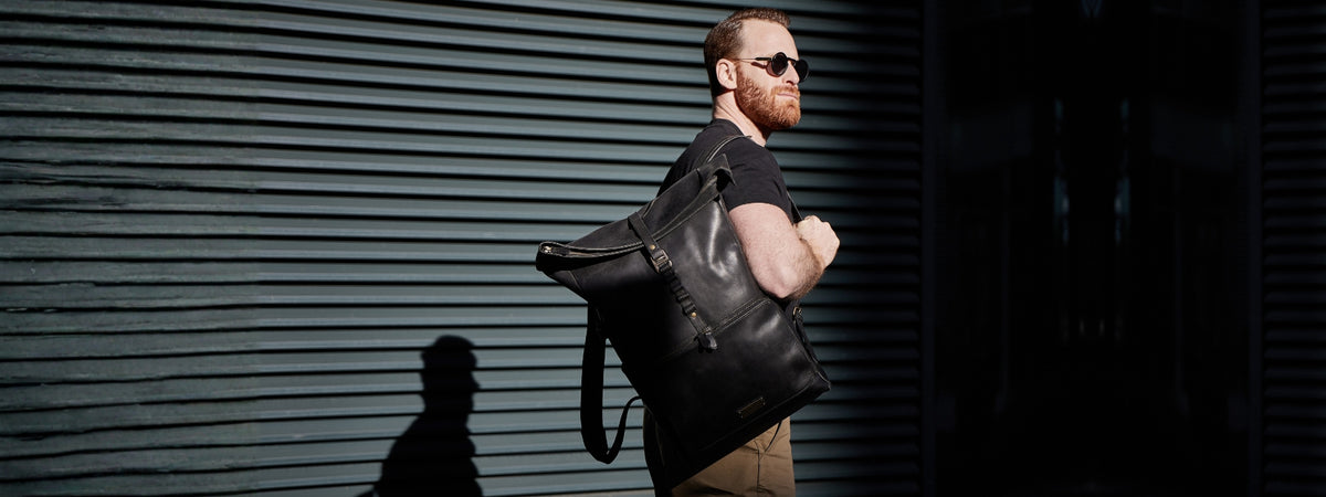 Homme portant un sac à dos rolltop en cuir noir sur une épaule devant une façade sombre