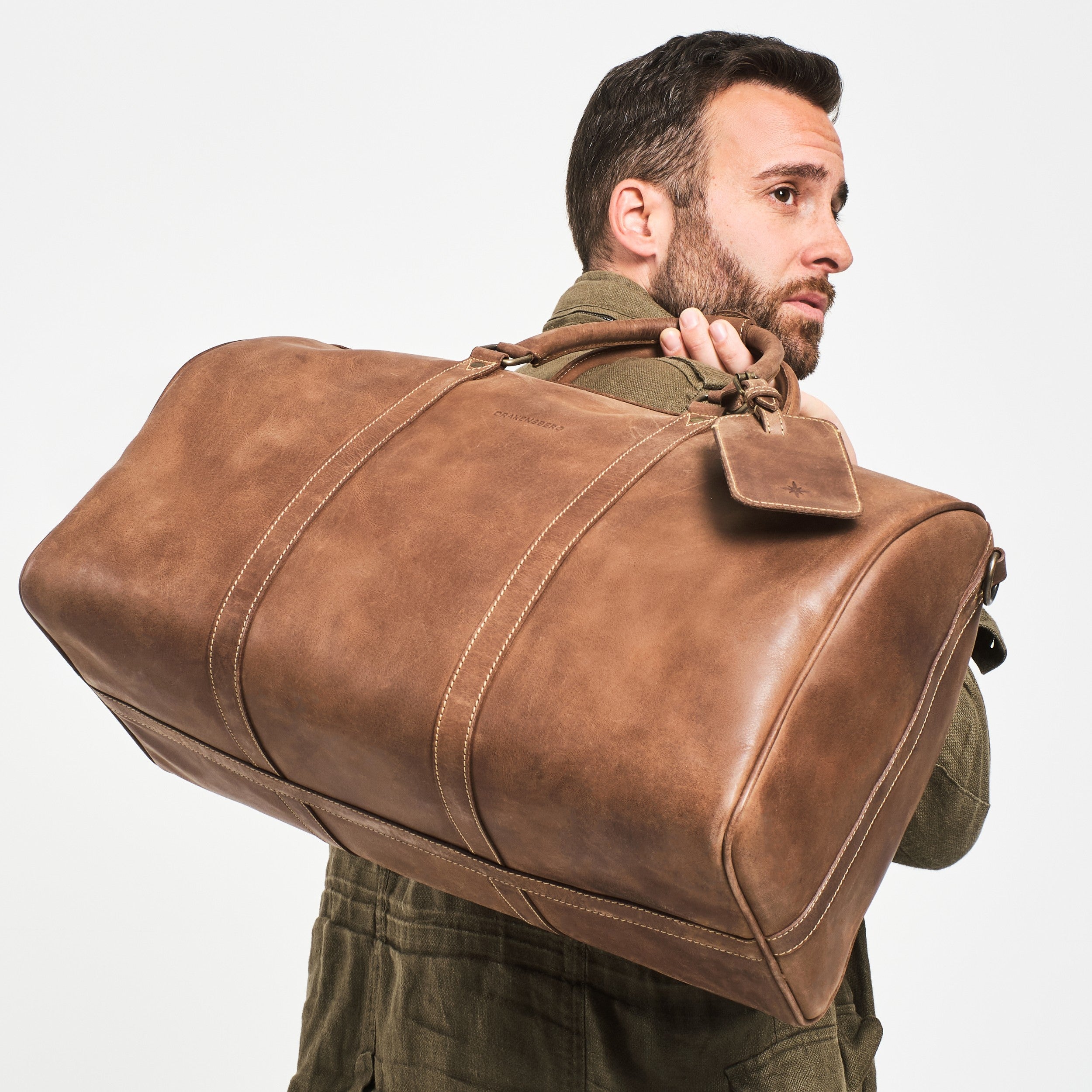 Dunkelhaariger Mann mit Weekender James 50 aus Leder für Herren und Damen in Havanna-Braun – handgefertigte Reisetasche aus Büffelleder im klassischen Vintage-Stil.