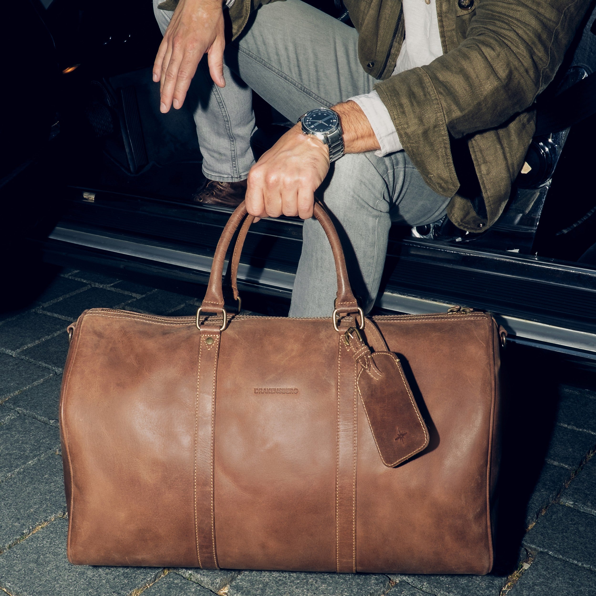 DRAKENSBERG Leder Weekender James 50 aus Leder für Herren und Damen in Havanna-Braun – handgefertigte Reisetasche - steht vor Autotür