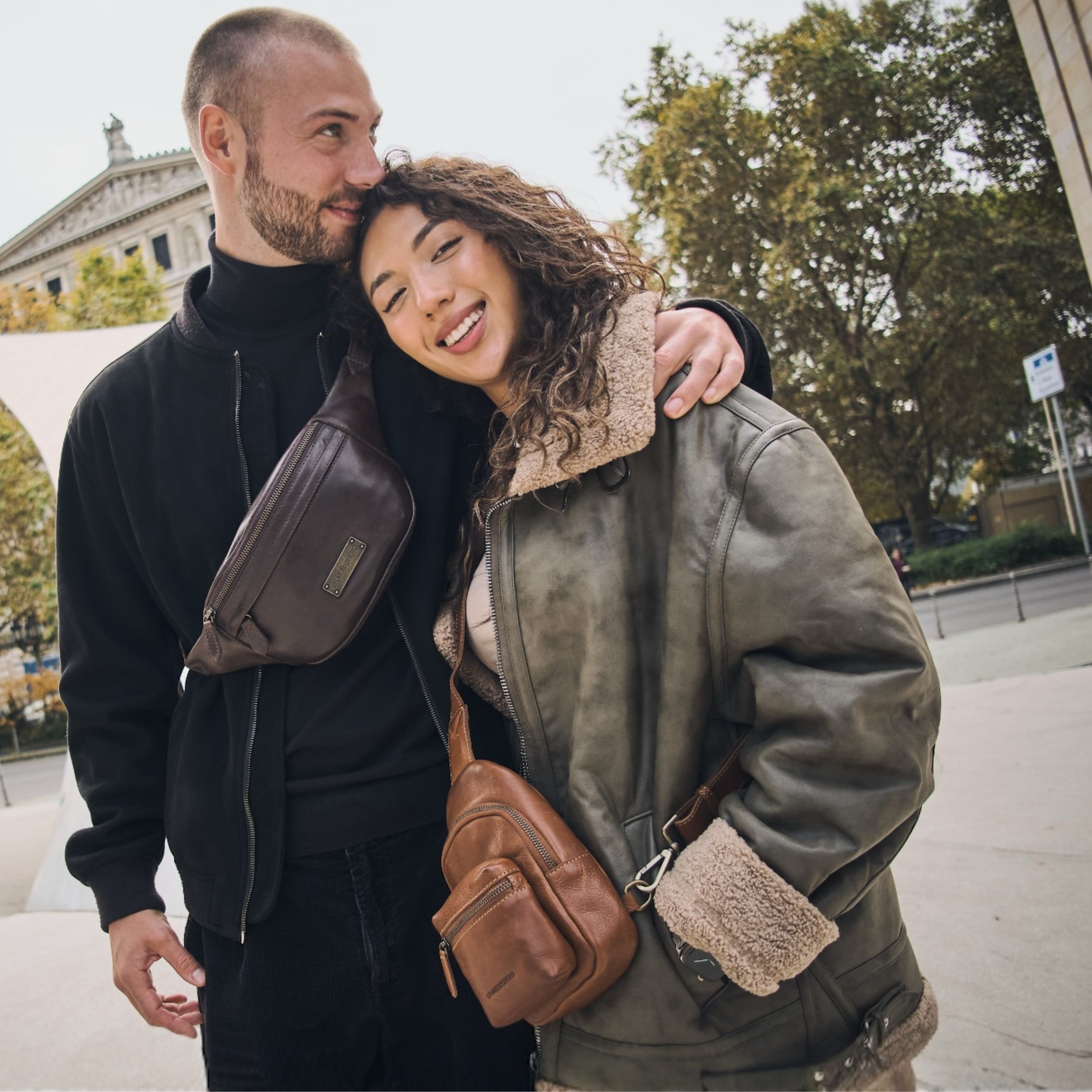 Ein junges Paar steht in Frankfurt vor der Oper, das weibliche asiatische Modell trägt eine Cross Bag Marlen in Karamell-Braun aus Leder