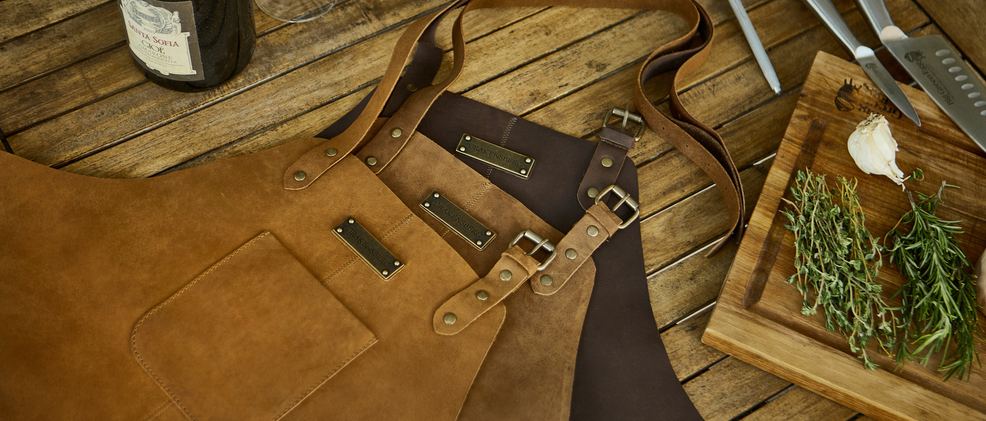 Hochwertige Grillschürzen aus Leder von DRAKENSBERG liegen auf einem Teakholz-Tisch, auf dem Tisch ist ein Schneidbrett, Kräuter und Messer zu sehen