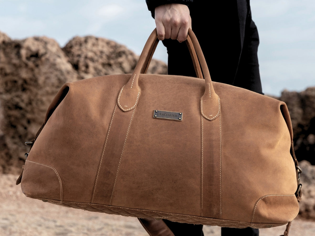 Sac de voyage en cuir marron tenu par les anses dans un paysage rocheux