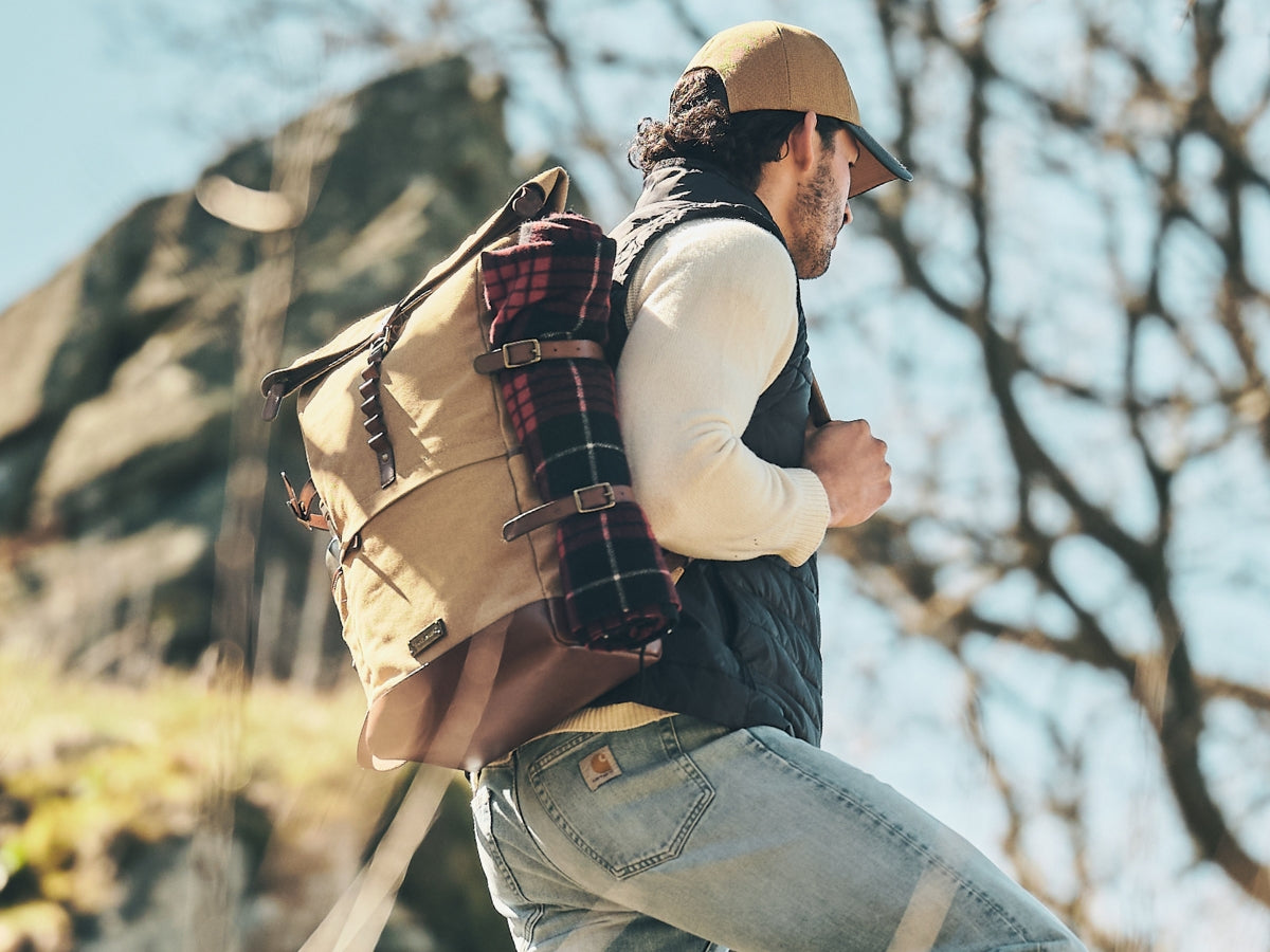 Homme portant un sac à dos rolltop en toile beige avec une couverture à carreaux attachée, dans un paysage rocheux