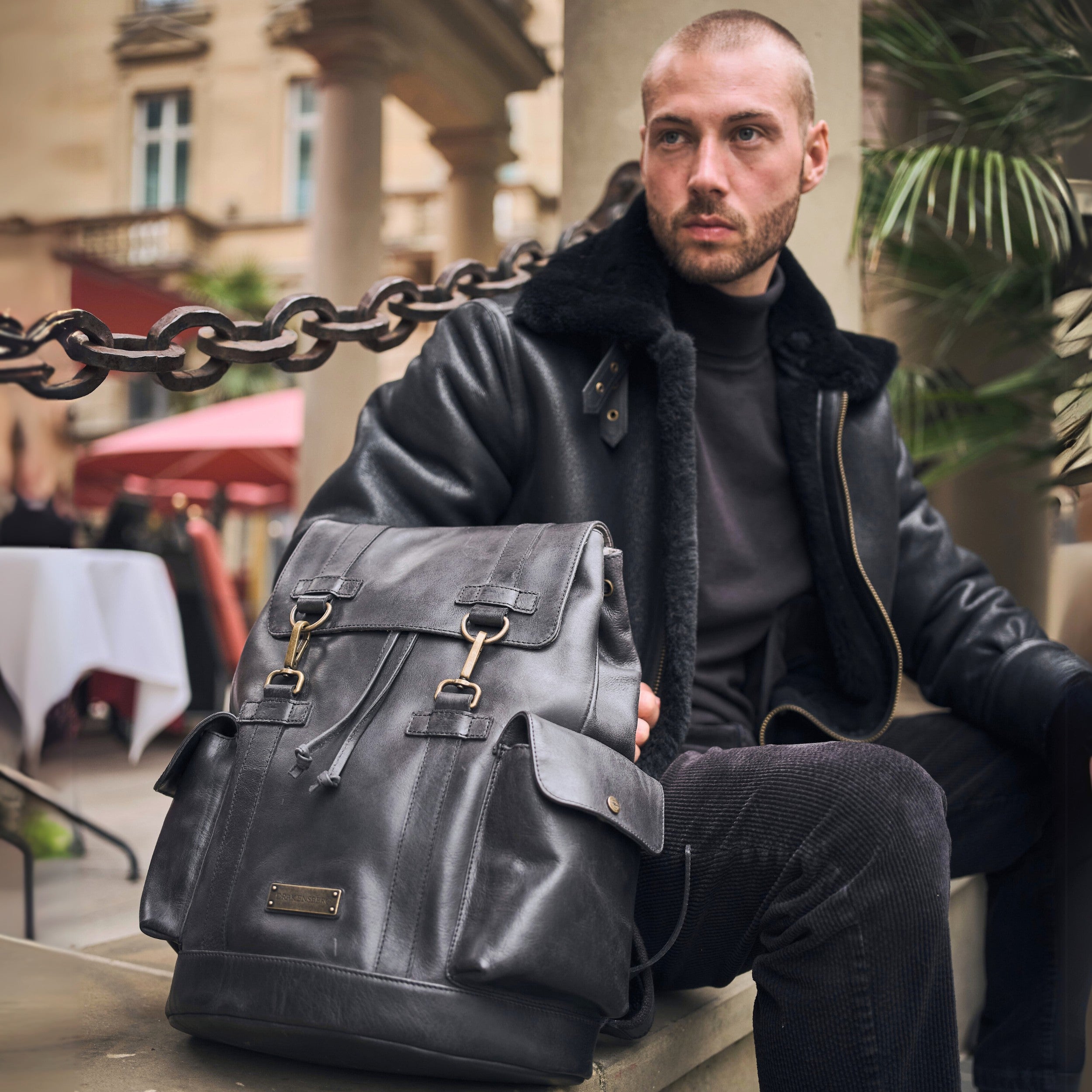 Männliches Modell sitzt auf einer Bank und hat einen schwarzen Rucksack Herren Leder vor sich stehen