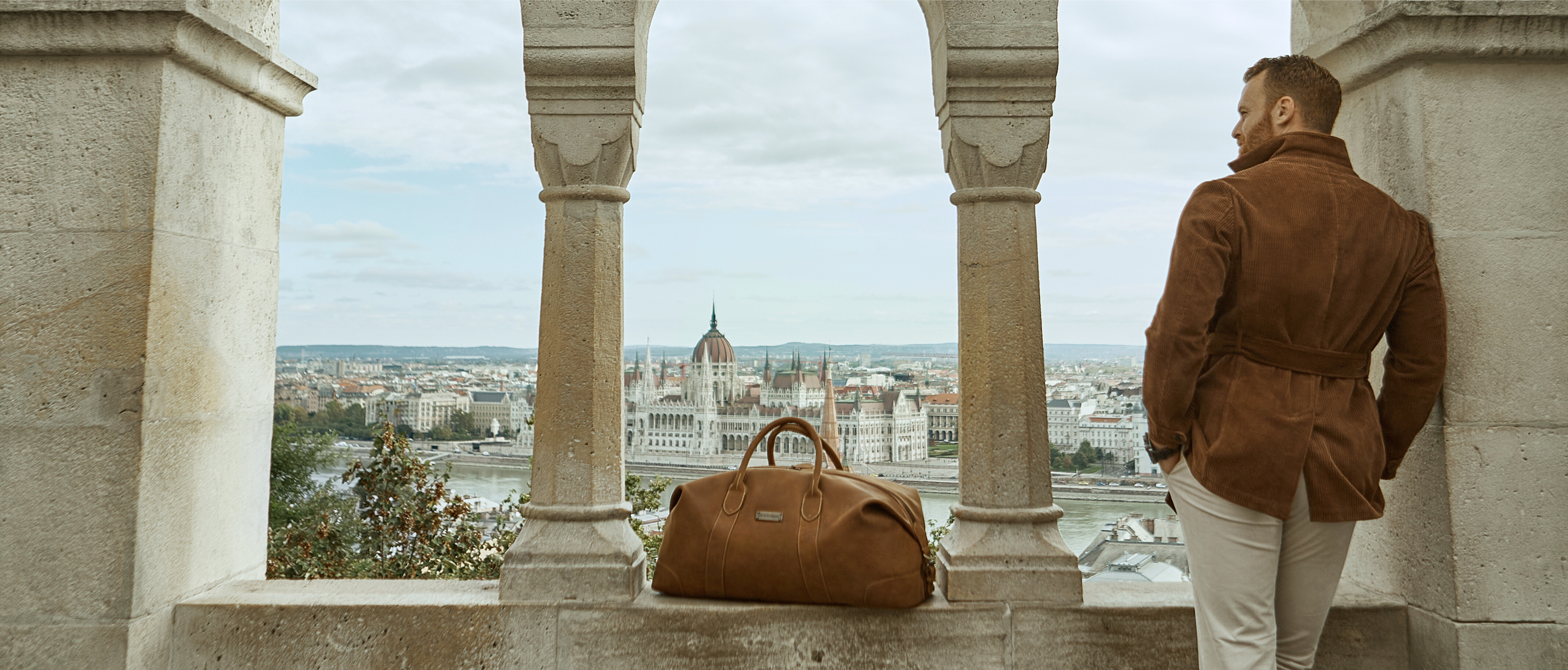 Mann steht auf einem Balkon und Blick auf Budapest, er hat eine Reisetasche aus Leder dabei-Pano