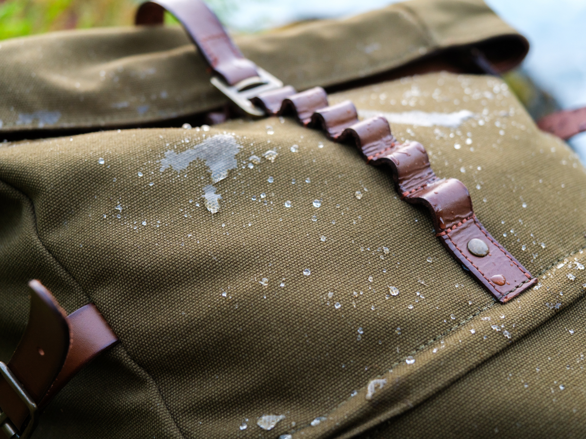 Water droplets beading on the canvas of the Wyatt backpack beside its leather closure strap.