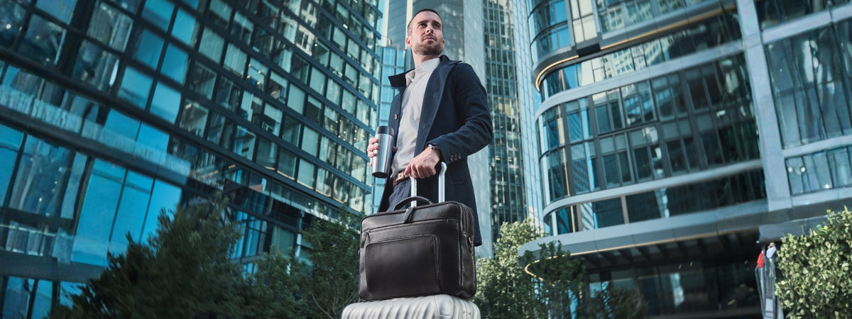 Mann mit schwarzer Leder-Laptoptasche und Rollkoffer vor modernen Bürogebäuden
