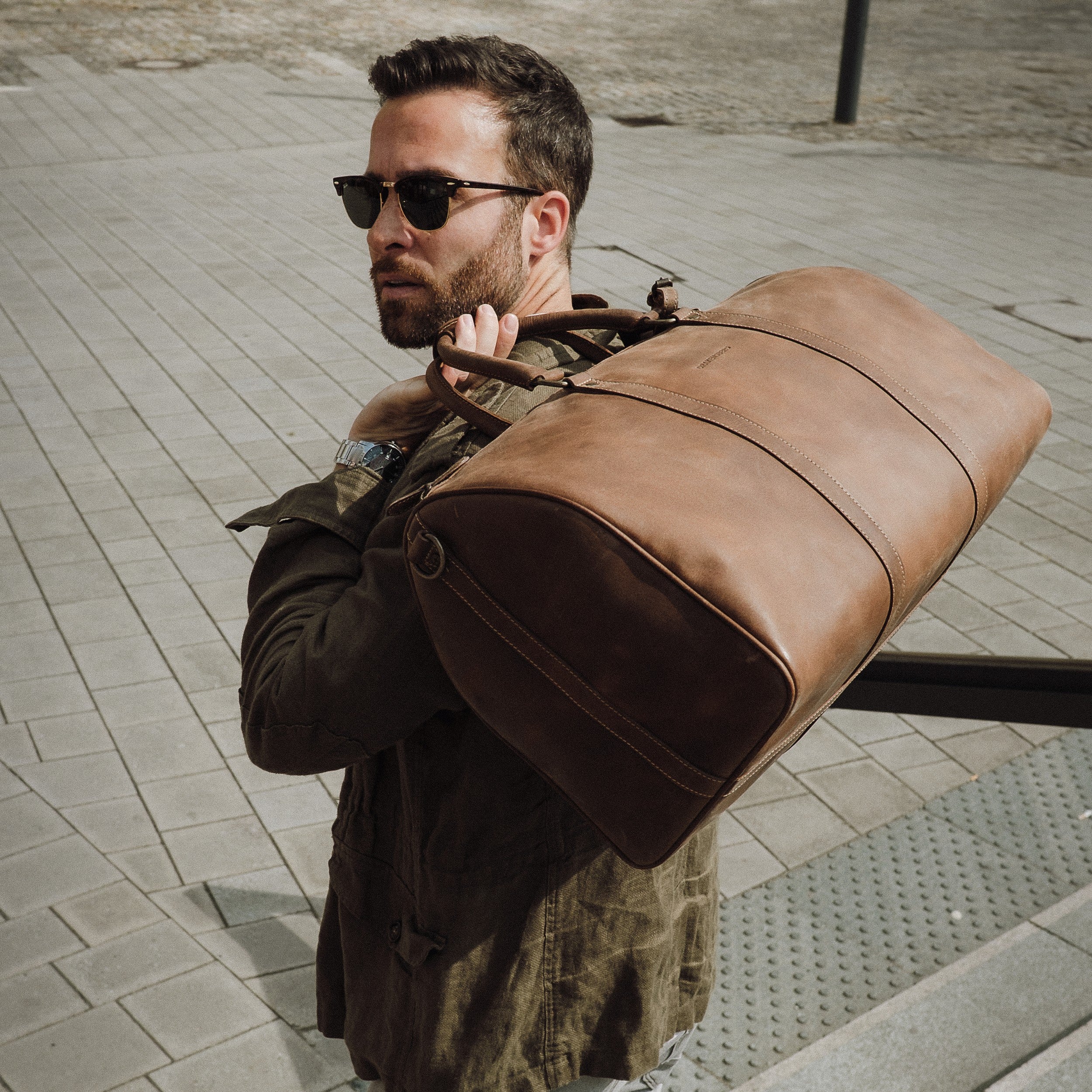 Dunkelhaariger Mann mit Sonnenbrille trägt Leder Weekender James 50 für Herren und Damen in Havanna-Braun – handgefertigte Reisetasche - Boardgepäck