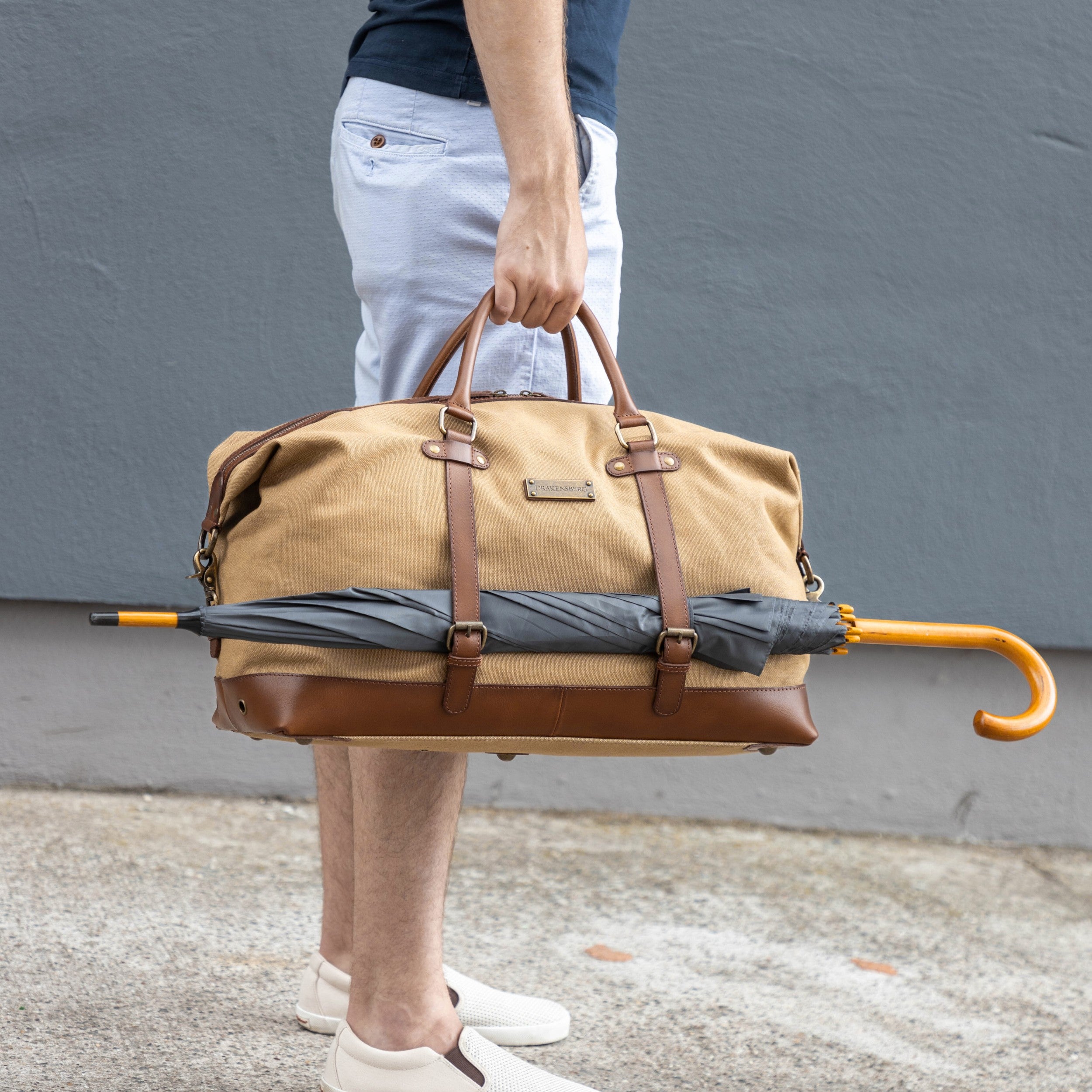 Canvas Weekender Owen Khaki-Beige mit Leder-Elementen - Mann trägt die Sporttasche befüllt und an der Seite ist ein großer Regenschirm befestigt
