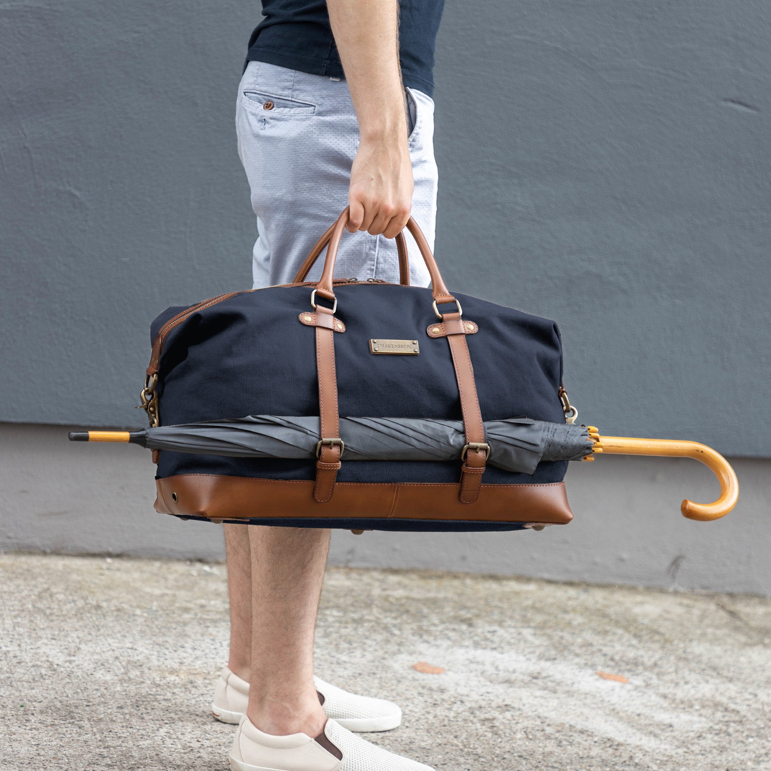 Canvas Weekender Owen Marine-Blau mit Leder-Elementen - Mann trägt die Sporttasche gefüllt und an der Seite ist ein großer Regenschirm befestigt