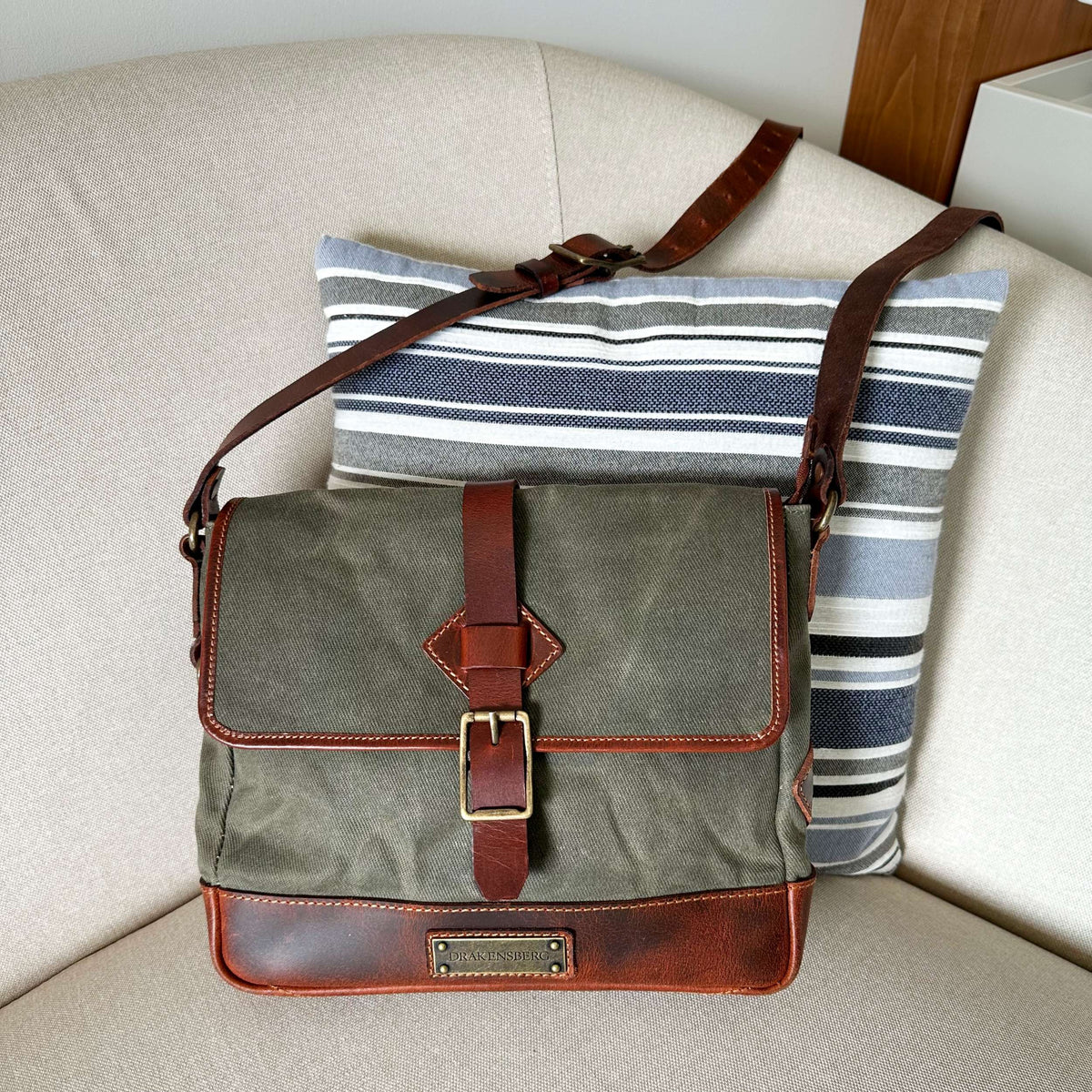 Sac bandoulière Nate en toile verte avec détails en cuir brun sur un fauteuil clair